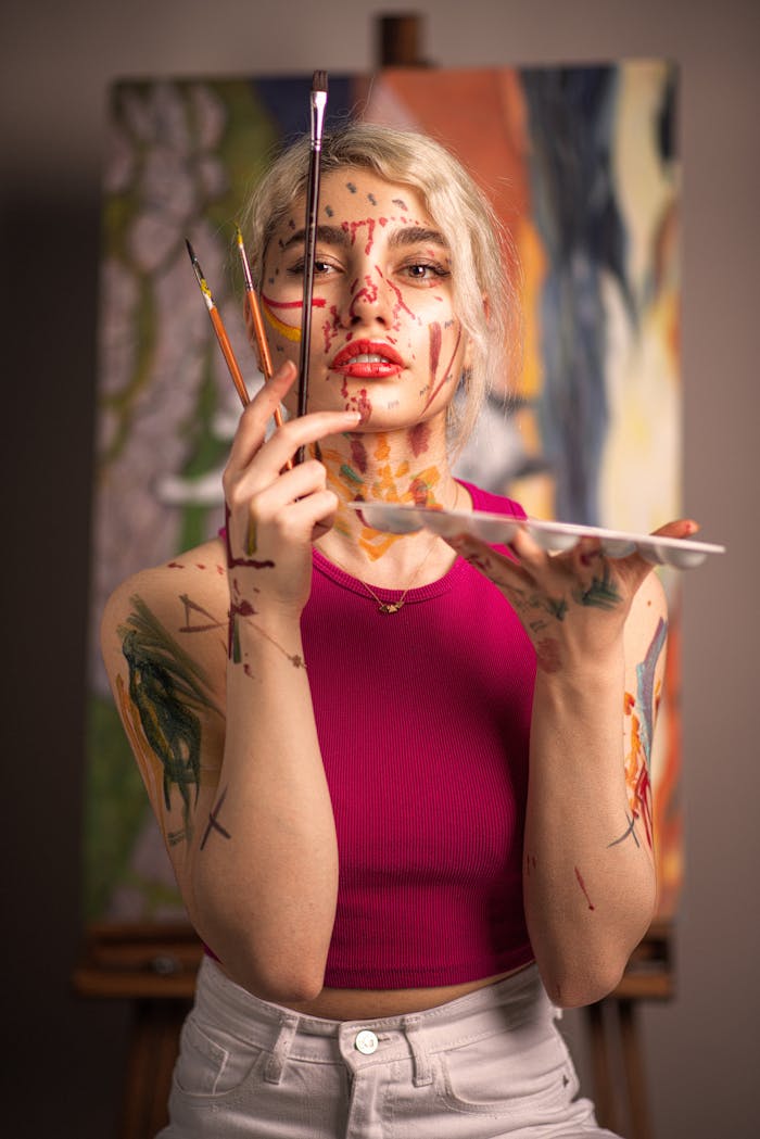 A young woman with paint on her face holding brushes in an art studio.